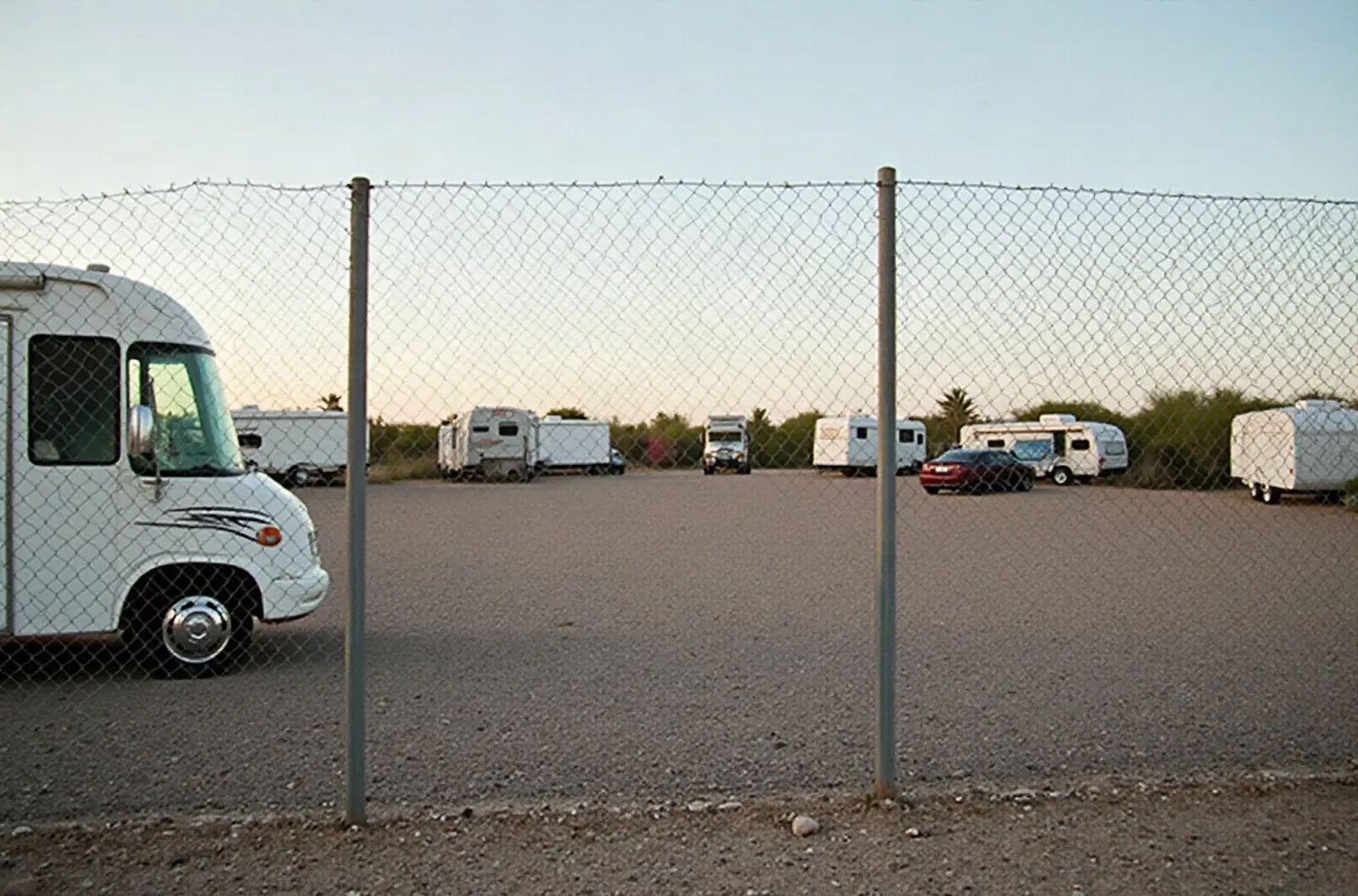 Chain-link fence corner of a gravel RV lot with a fifth wheel in the distance, illustrating a budget-friendly RV storage spot in the desert.