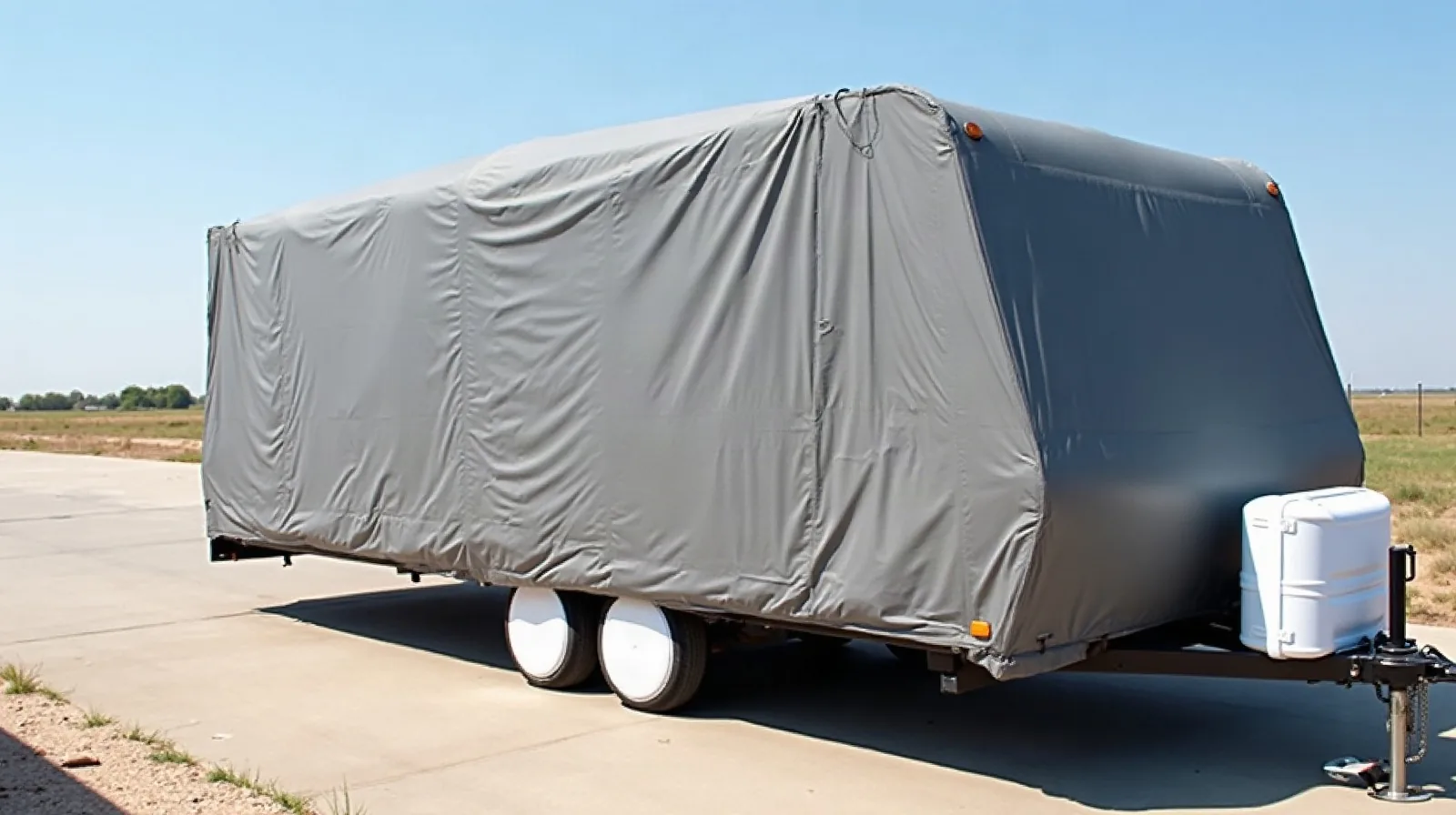 Travel trailer covered with gray breathable RV storage cover, straps underneath, white tire covers, parked on open Texas lot with wind rippling fabric